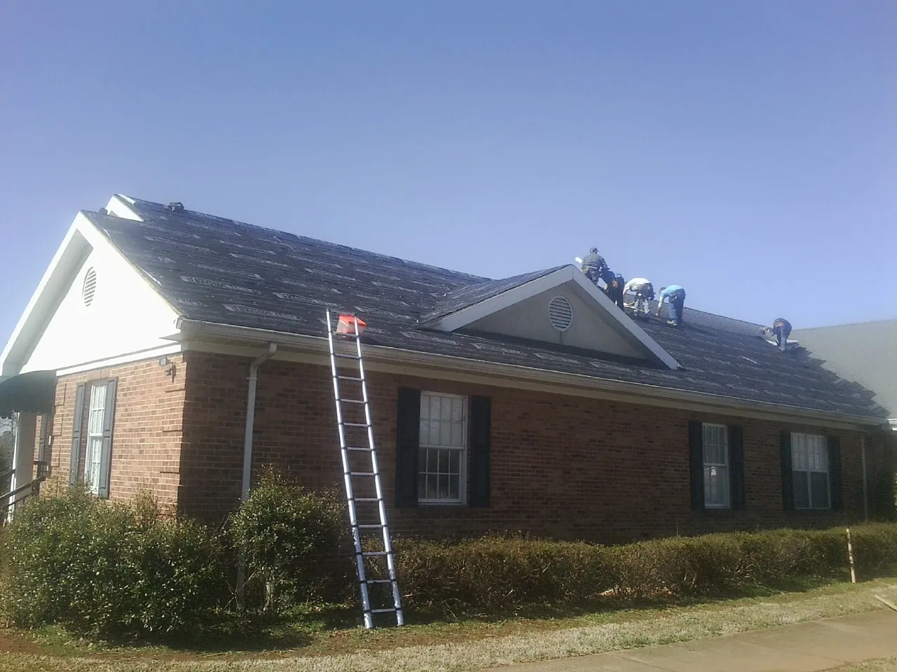 Crew tearing shingles off a brick ranch roof with a ladder set — Metal Roof Repair work by our crew in Howard City