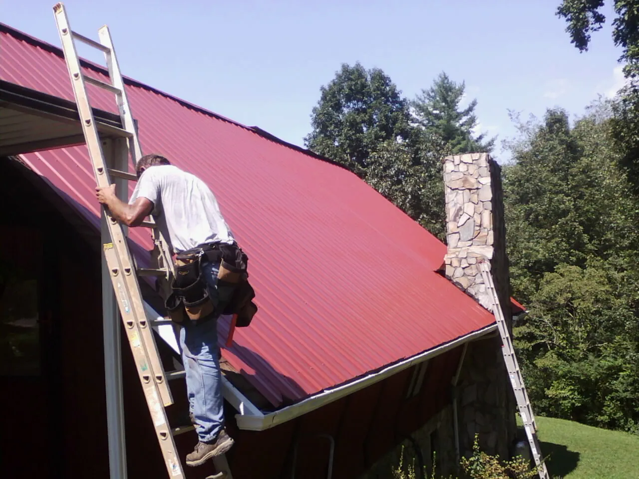 Roofer on a ladder beside a red metal roof and stone chimney — Soffit & Fascia Repair by Roofing Brothers in Howard City